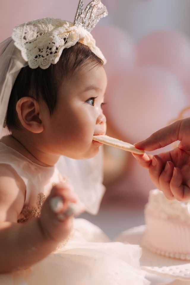 Baby girl in lace headband eating cookie at first birthday cake smash party with pink balloons.