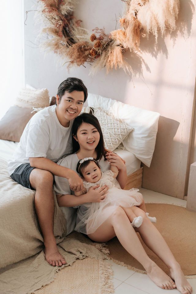 Happy Asian family of three posing together on bedroom floor with boho dried floral wreath decor.
