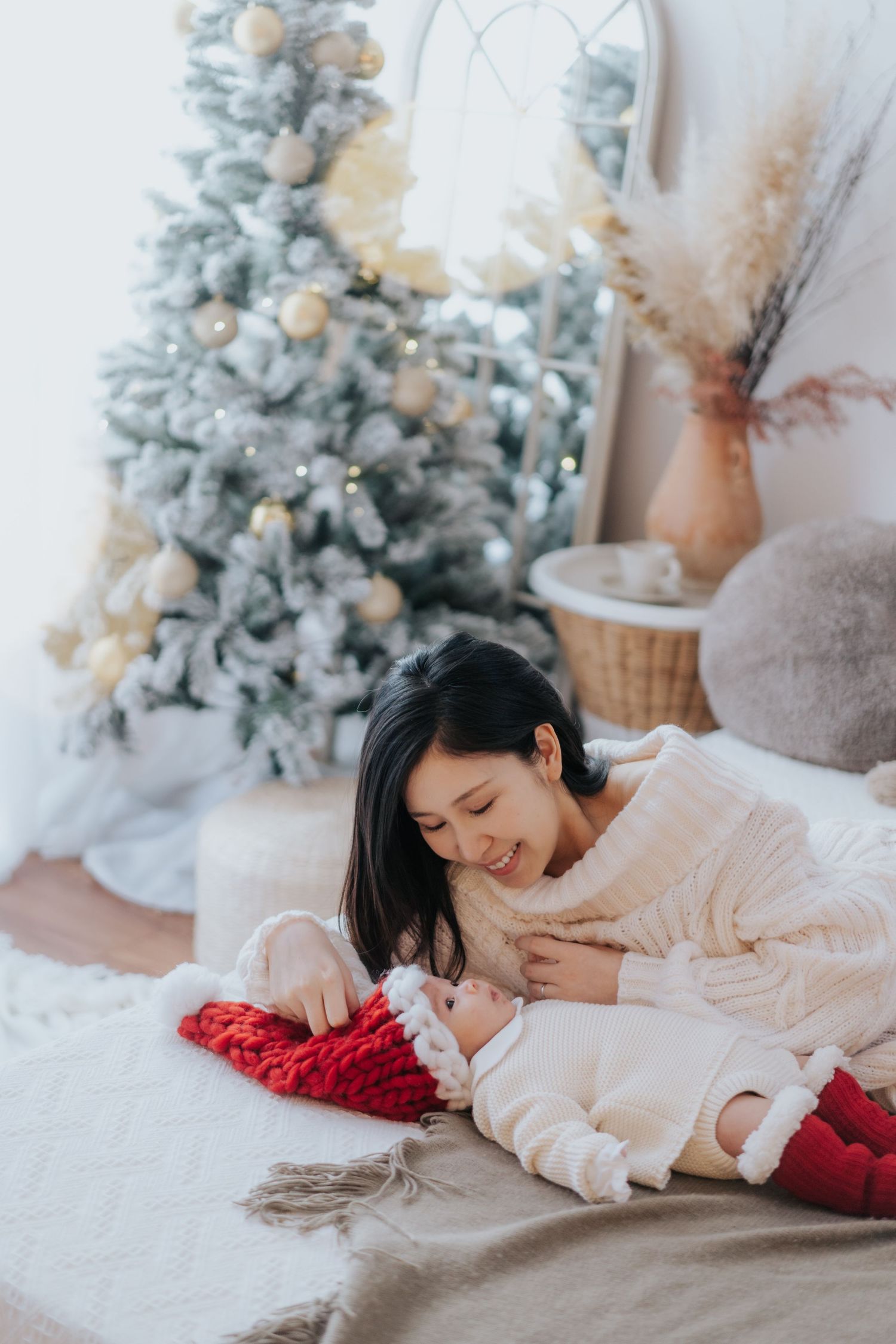 A mother tenderly holds her newborn in a Santa hat beside a decorated Christmas tree in a cozy white room.