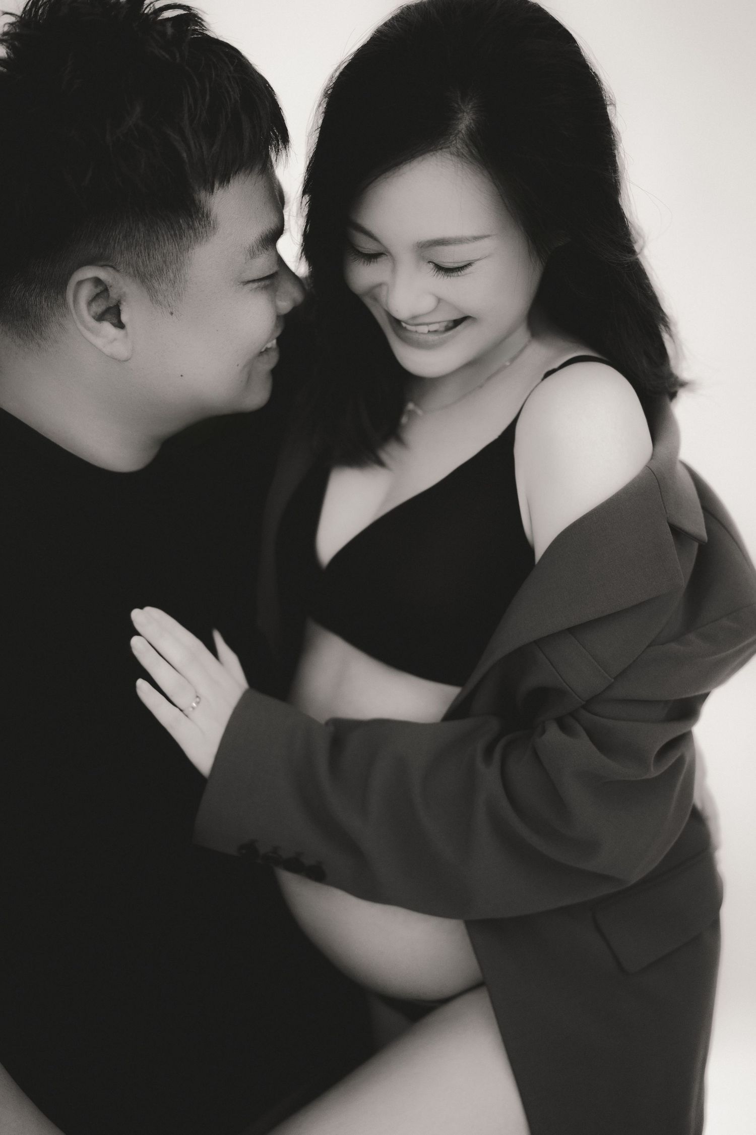 A black and white intimate maternity photo showing a couple embracing in a tender moment against a light background.