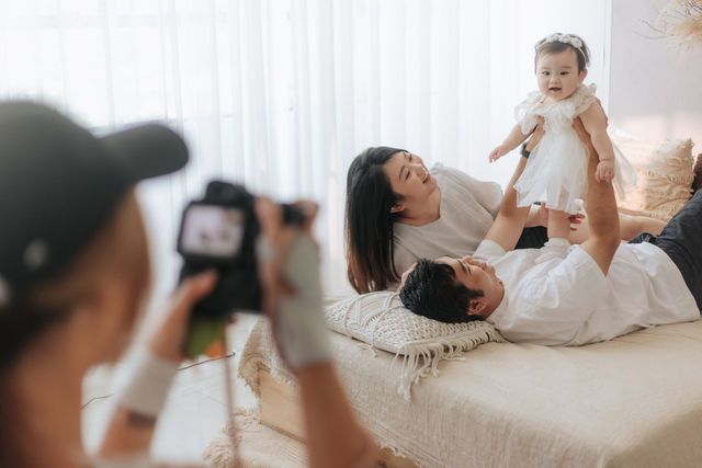 Photographer capturing happy family with baby girl being lifted on cozy bed indoors.
