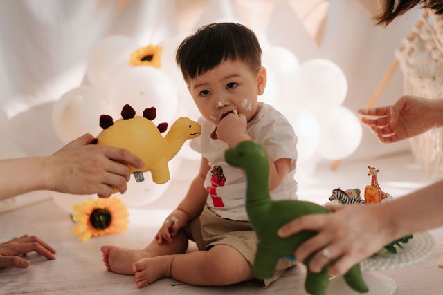 Cute toddler boy playing with colorful dinosaur toys surrounded by balloons and sunflowers in a bright, cheerful setting.