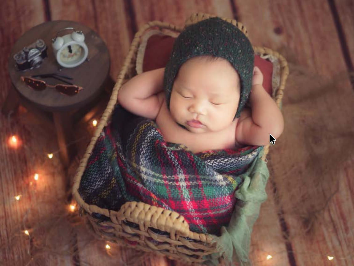Newborn baby sleeping peacefully in a wicker basket with plaid blanket, fairy lights, and vintage props on wooden floor.新生嬰兒安詳地睡在鋪著格子毯、掛著彩燈