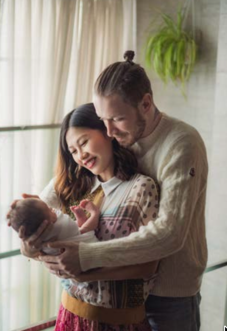 A couple shares a tender moment with their newborn baby by a window with a hanging plant in the background一對夫婦抱著剛出生的寶寶，在窗邊享受溫馨的時刻，窗外懸掛著一盆植物。