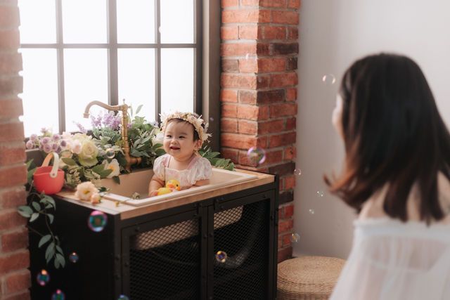 A joyful baby wearing a floral crown sits on a cabinet while bubbles float around as a woman watches nearby.