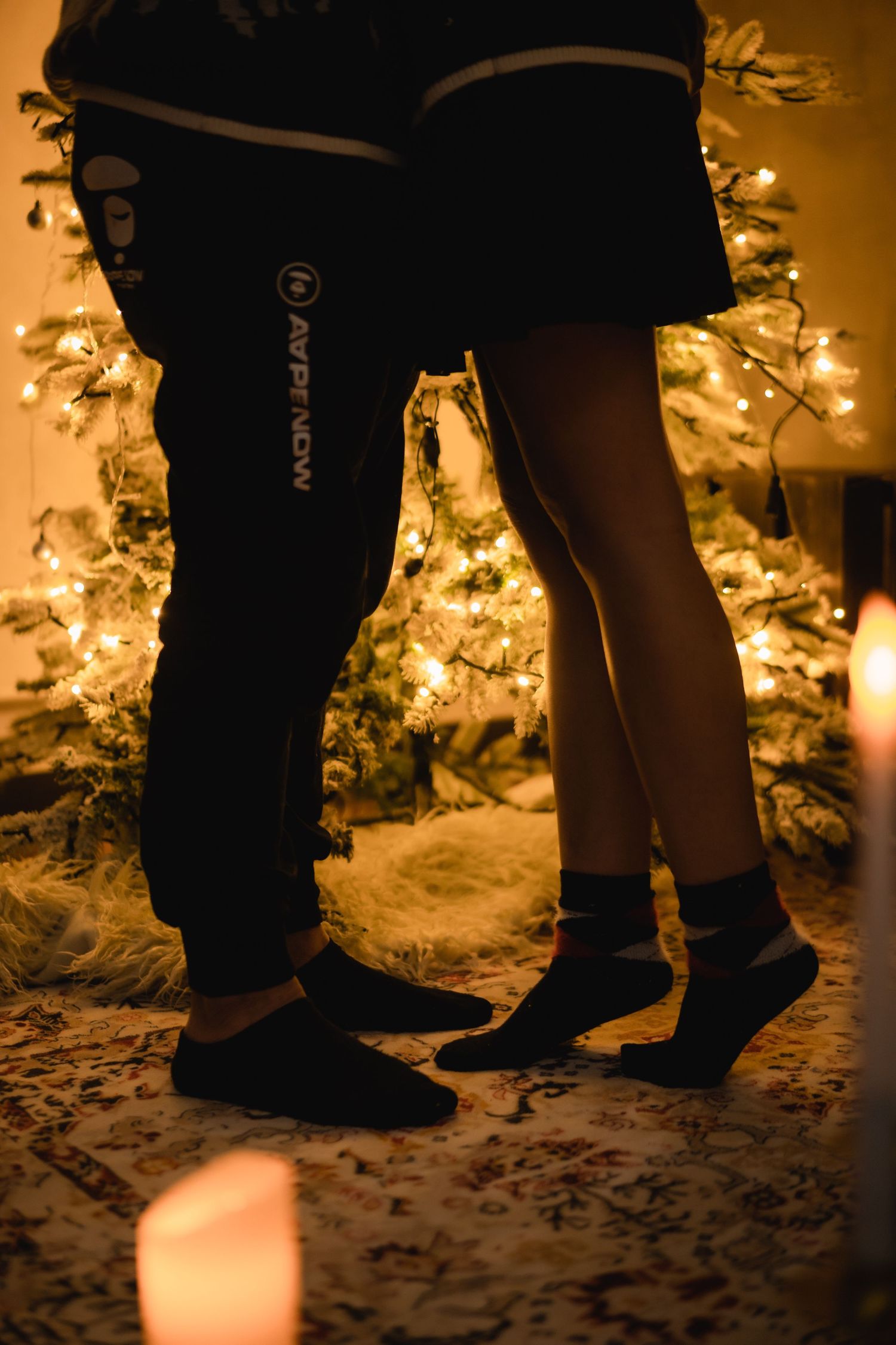 Two people standing by a glowing Christmas tree with candles, wearing dark pants and socks on a cozy rug.