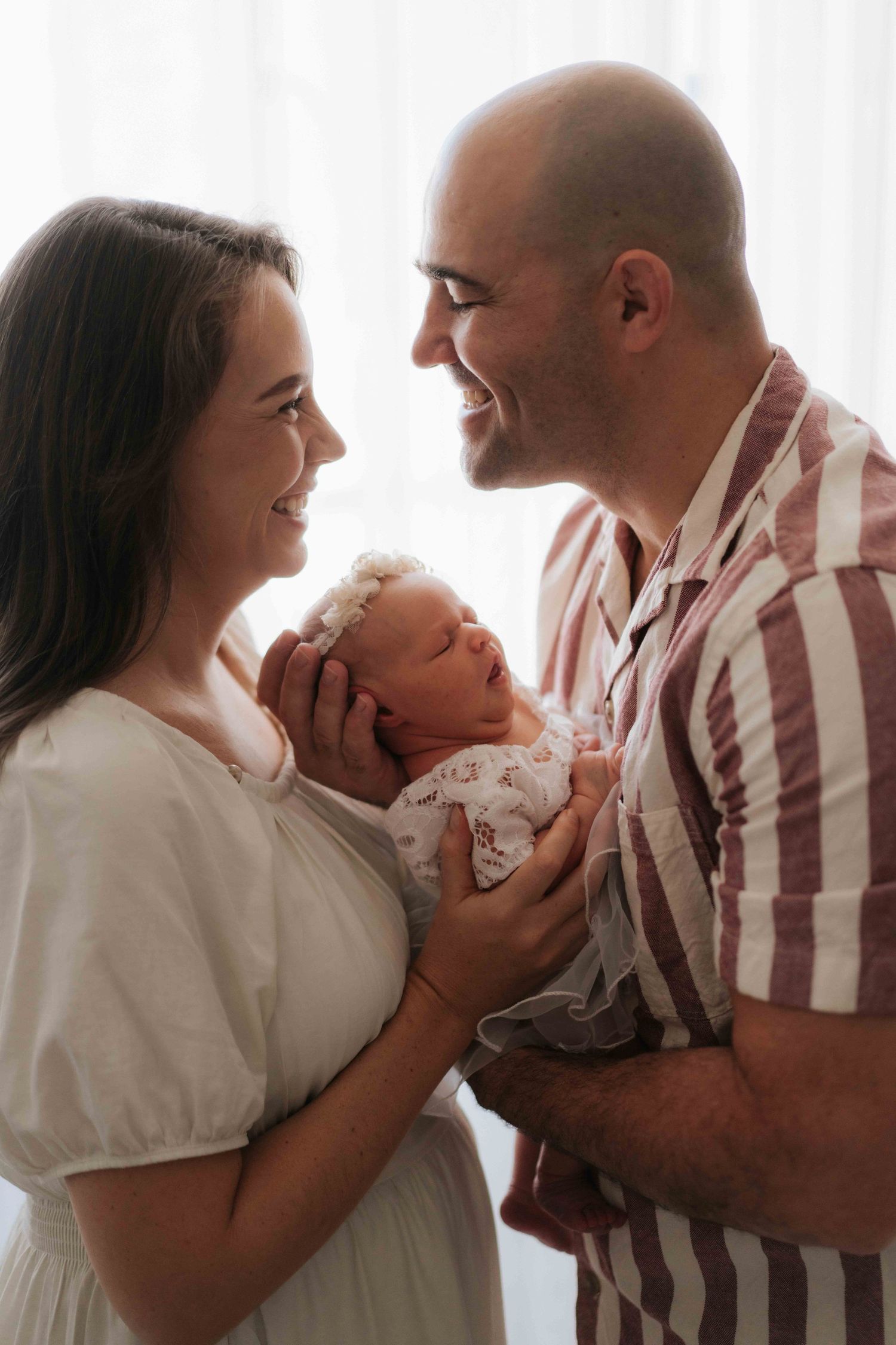 A couple shares a tender moment while holding their newborn baby against a bright backlit background.