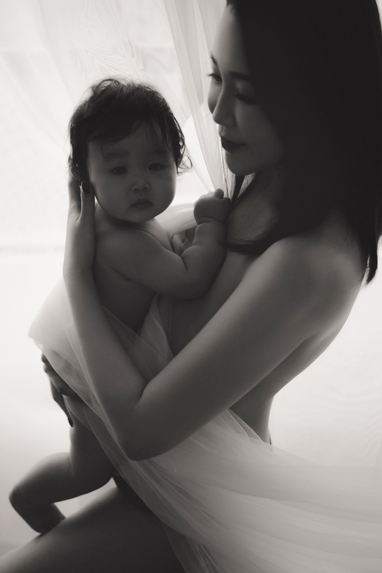 Black and white photo of a mother tenderly holding her baby near a bright window, wrapped in soft white fabric.一張黑白照片，照片中一位母親溫柔地抱著裹在柔軟白色布料