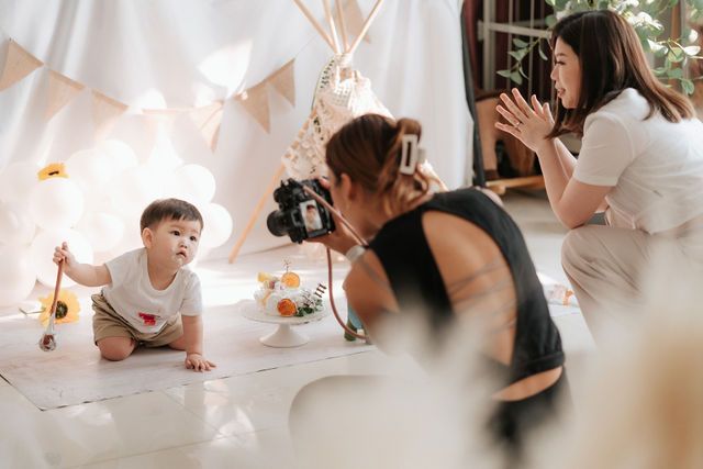 Two women photograph a cute baby crawling on a white mat in a decorated studio with a teepee backdrop.