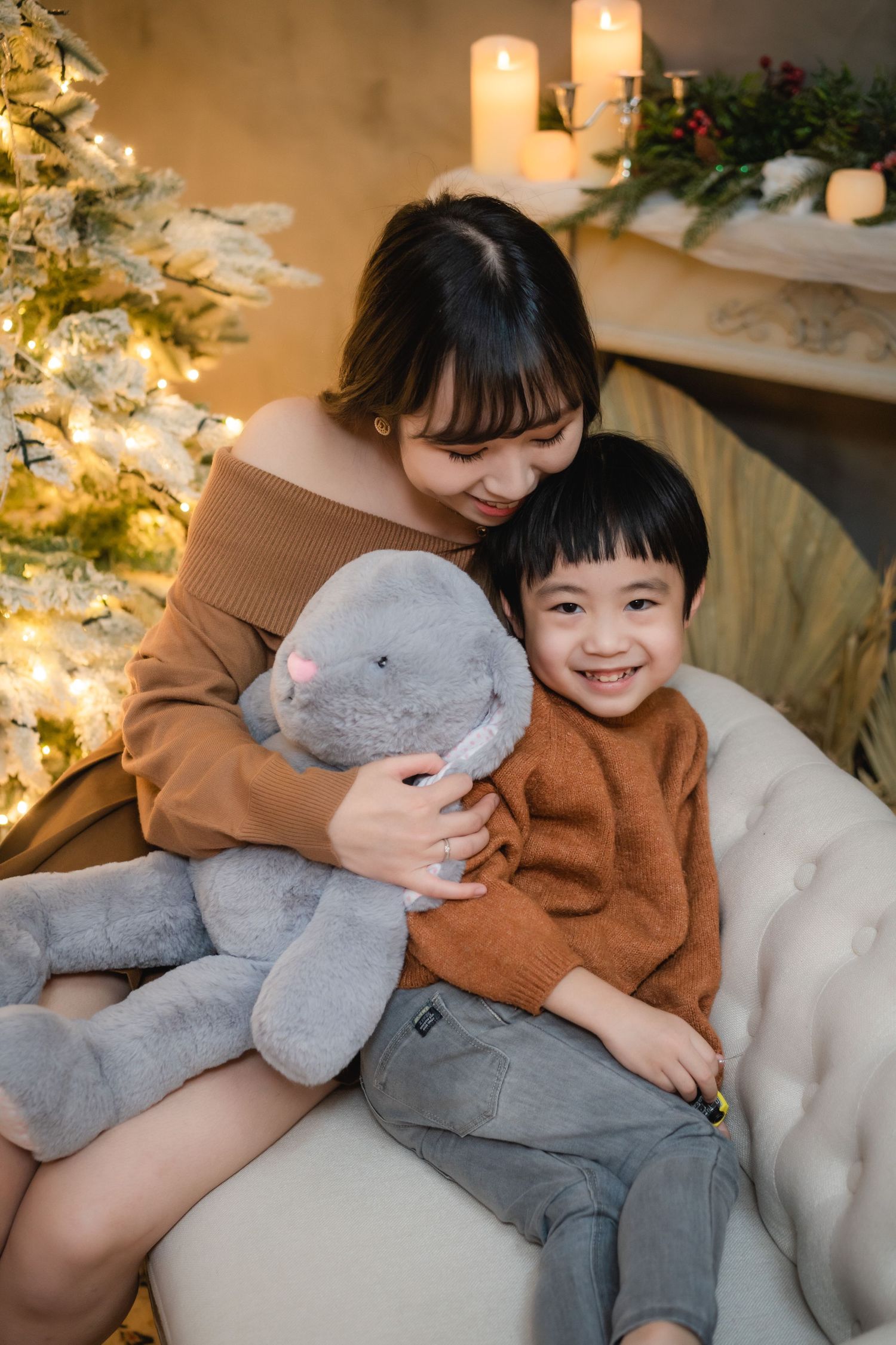 Asian mother and son in brown sweaters sitting on sofa with stuffed bunny near Christmas tree and fireplace.