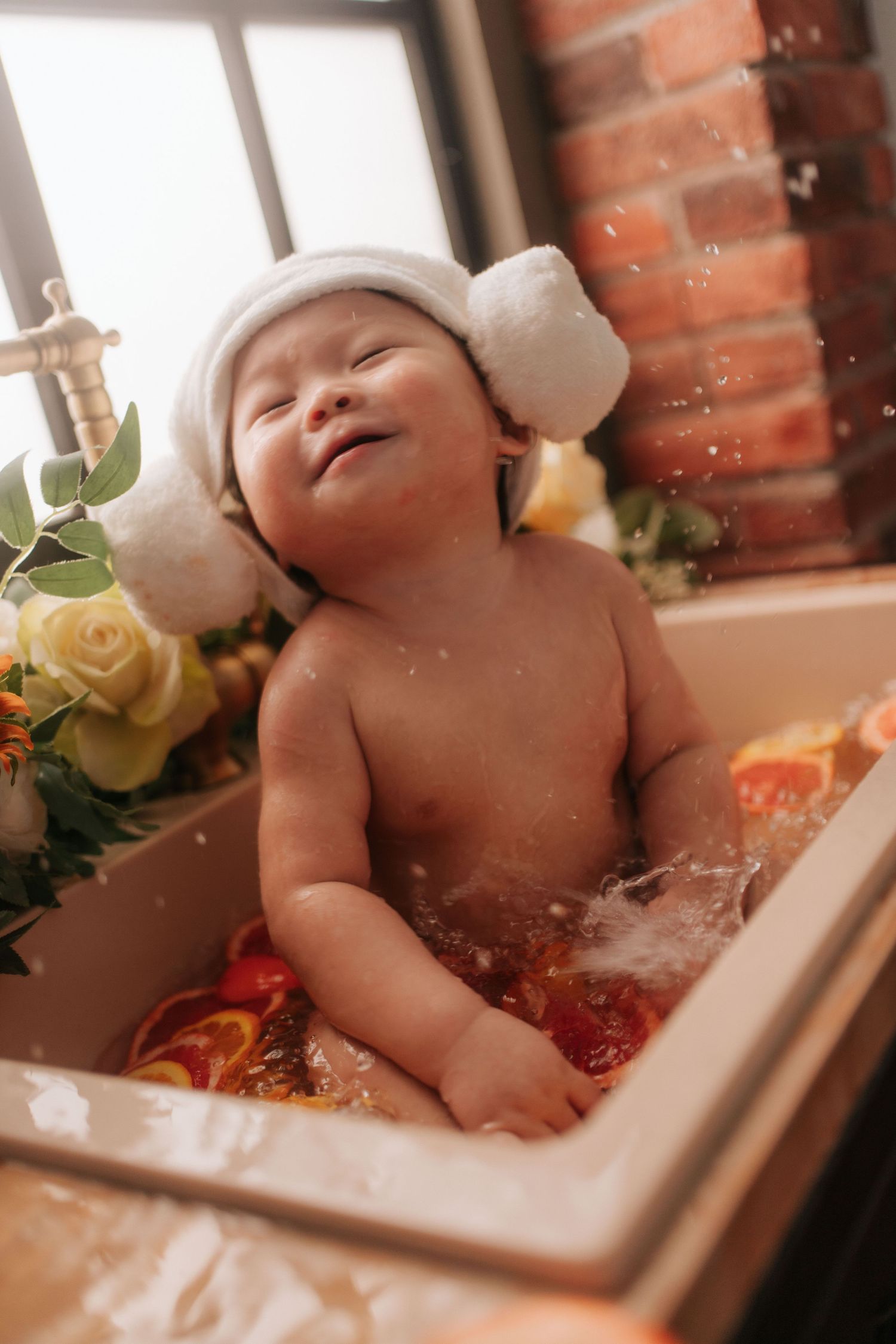 Smiling baby in a white hat sits in a sink bath with citrus slices and flowers near a brick wall.一個戴著白色帽子的微笑嬰兒坐在水槽浴缸裡，旁邊有柑橘片和鮮花，靠近一面磚牆。