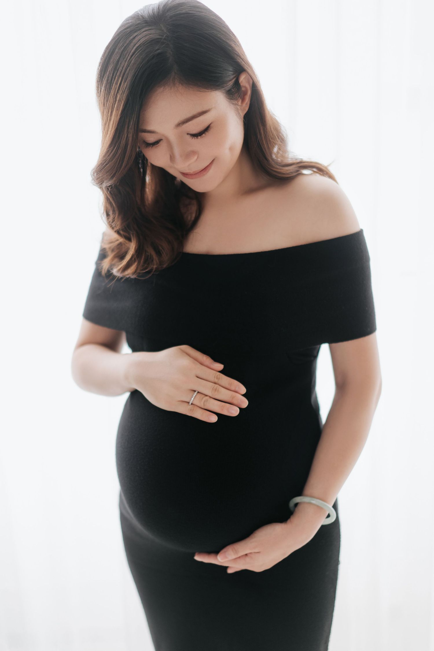 Maternity photo series showing woman in black off-shoulder dress cradling baby bump against white background.一組孕婦寫真，照片中一位身穿黑色露肩連身裙的女子在白色背景前