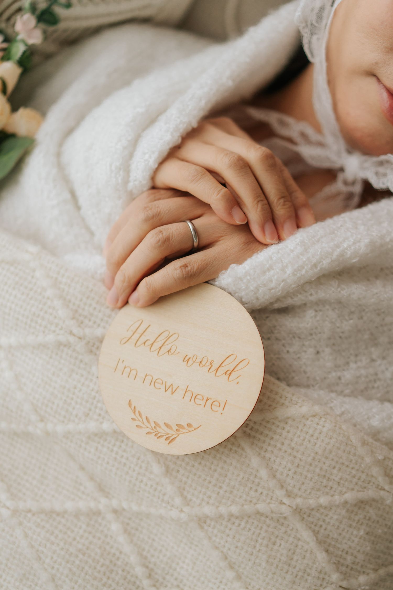 Newborn baby resting on white knit blanket beside a wooden Hello World I'm New Here milestone disc.