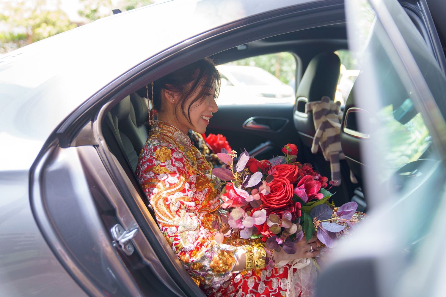 A bride in a floral red dress sits in a car holding a vibrant bouquet of pink and red roses on her wedding day.