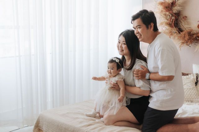 Happy Asian family with parents and toddler daughter sitting on bed near white curtains in bright room.