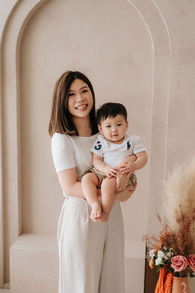 Asian mother smiling while holding her toddler son in a bright studio with floral decor.