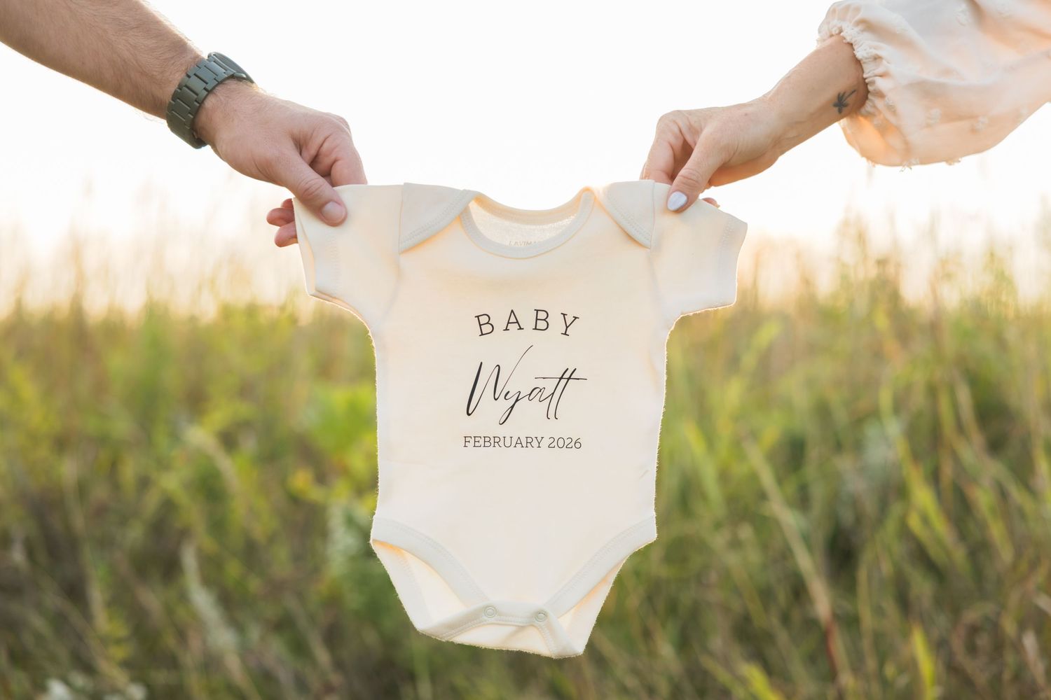 Hands holding up white baby onesie with custom text against sunset field background.