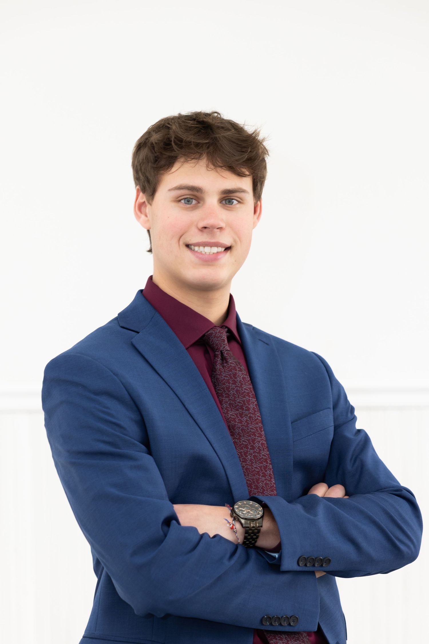 Professional portrait of someone in a navy blue suit with burgundy shirt and tie standing with arms crossed.