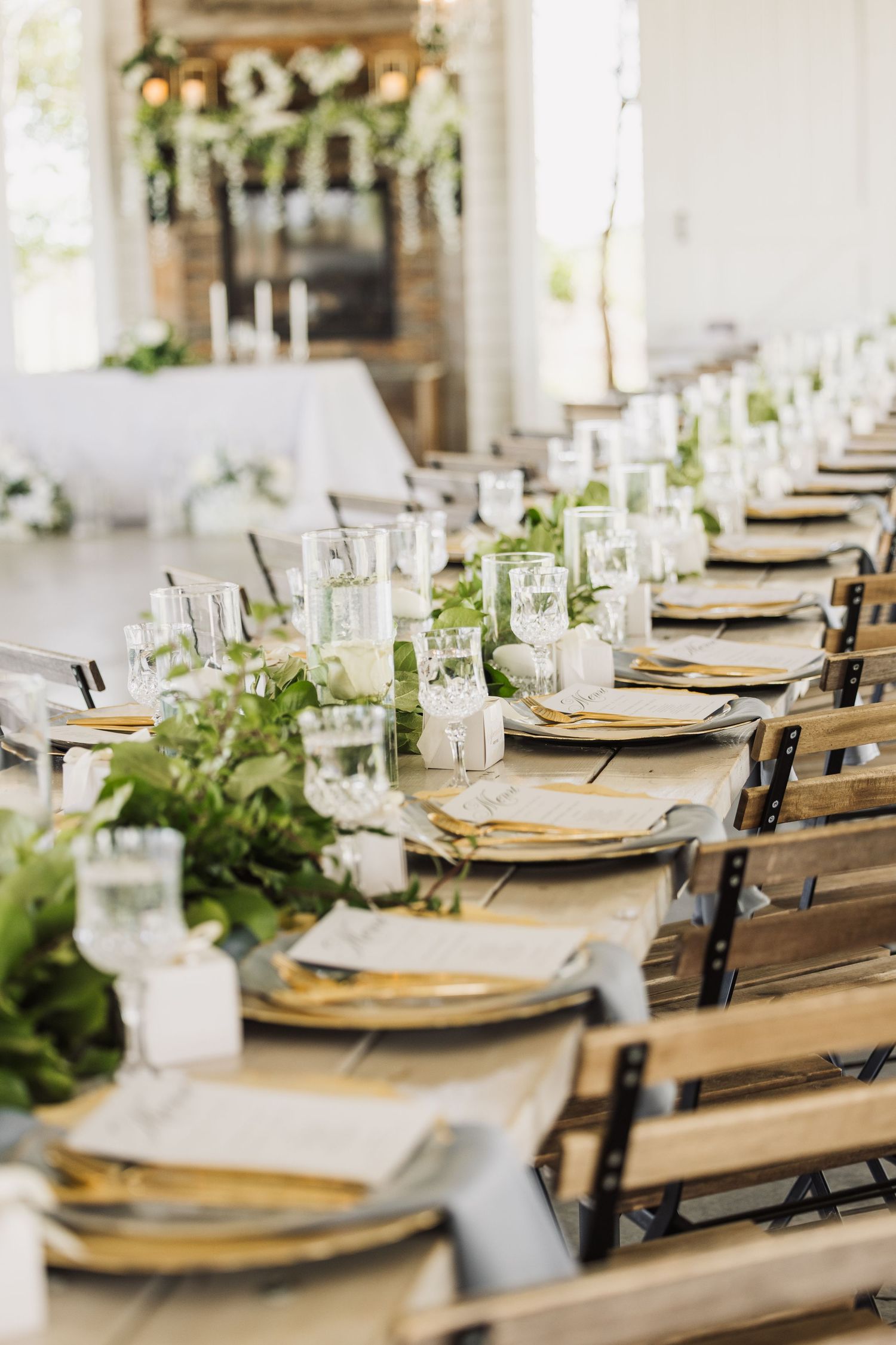 Rustic wedding reception table with gold chargers, greenery runners and wooden chairs.