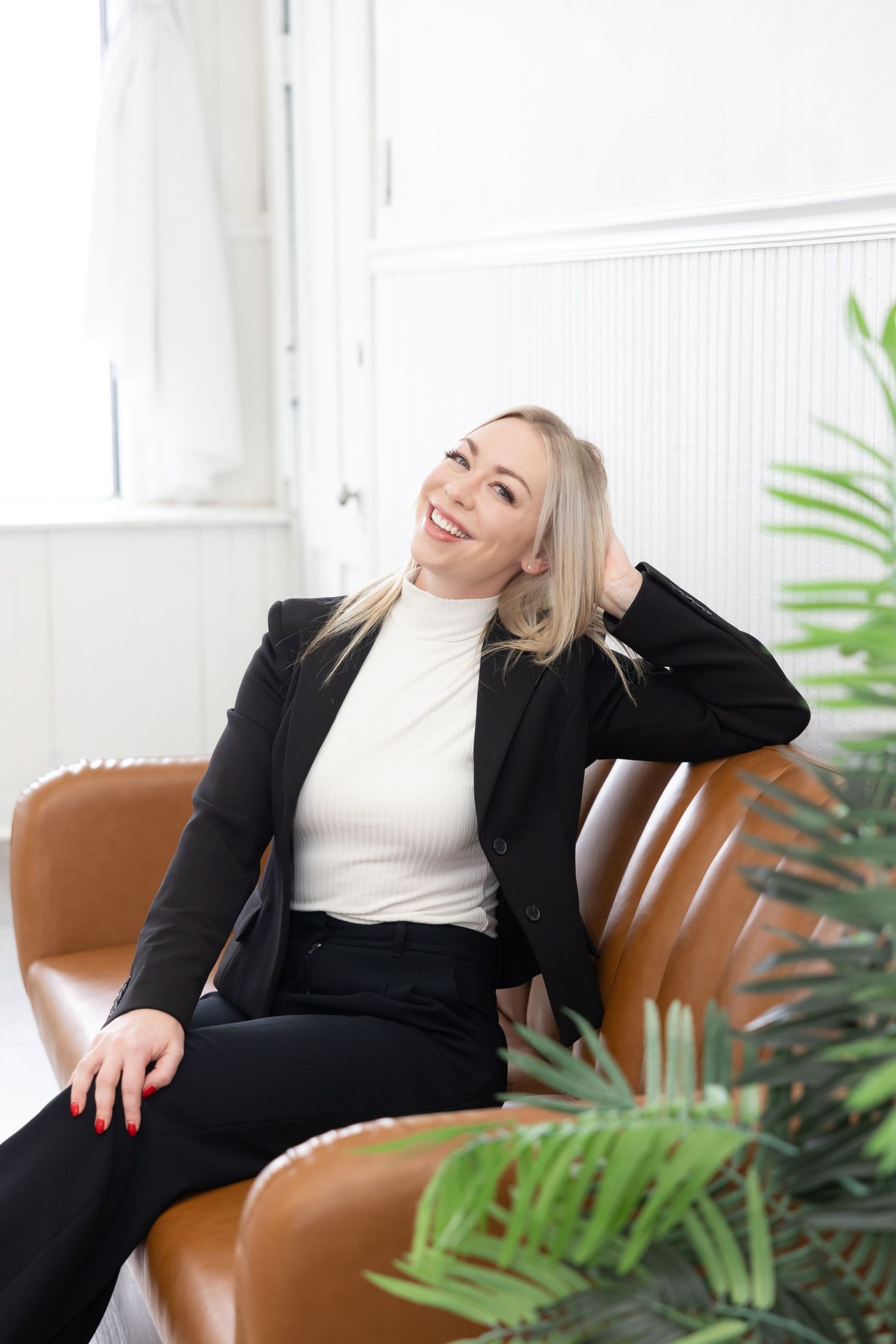 Professional in a black blazer and white turtleneck relaxes in a tan leather chair next to indoor plants.