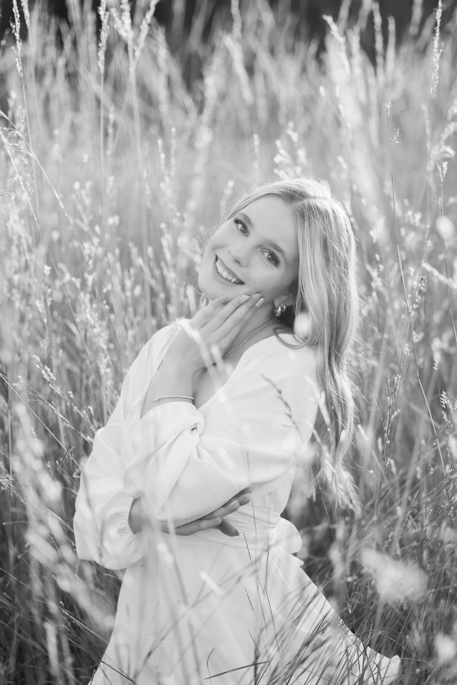 Black and white portrait of a smiling blonde woman in a white dress surrounded by tall wild grass.