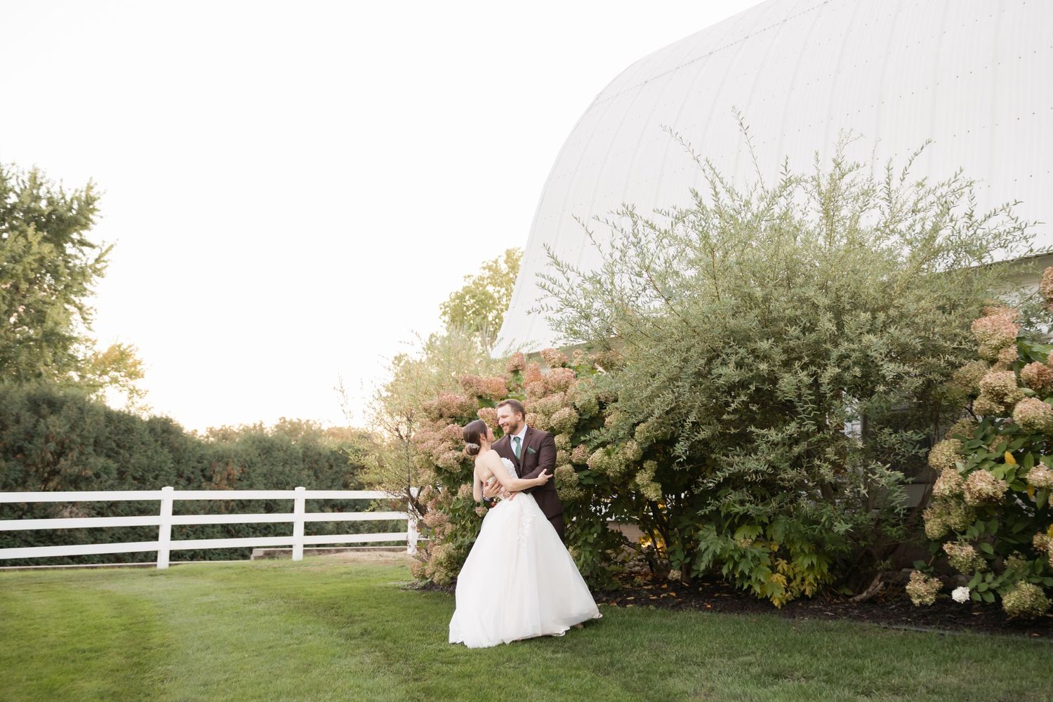 White barn and fence surrounded by blooming bushes on a scenic rural property.