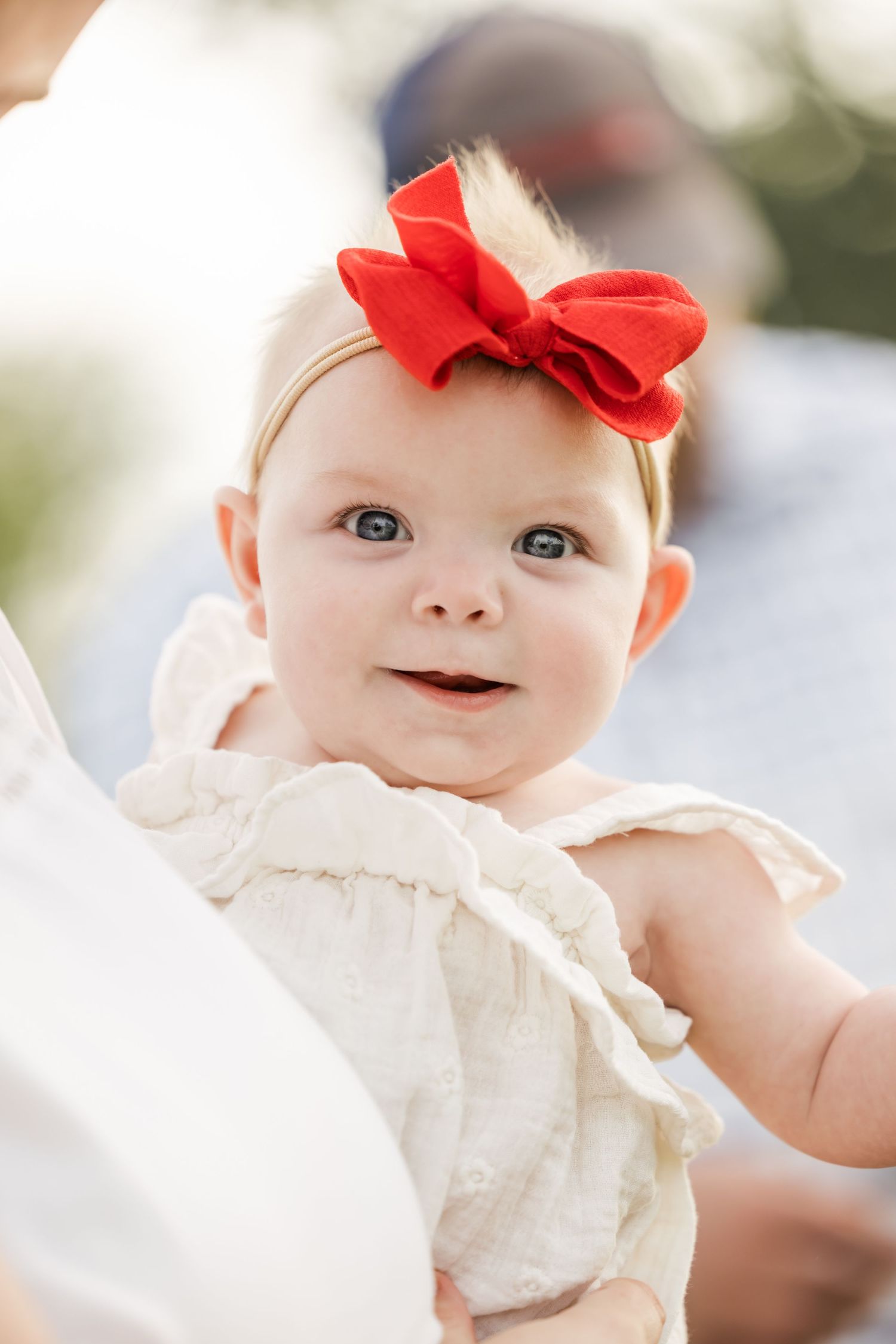 Baby wearing a red bow headband and cream-colored outfit smiles at camera.