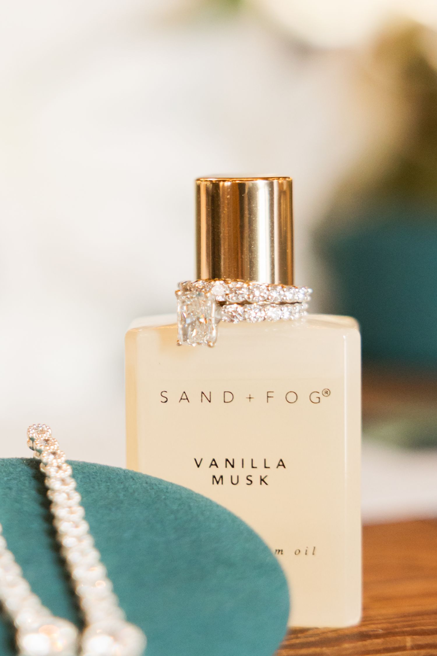 Sand + Fog vanilla musk perfume bottle with pearl necklace on teal surface.