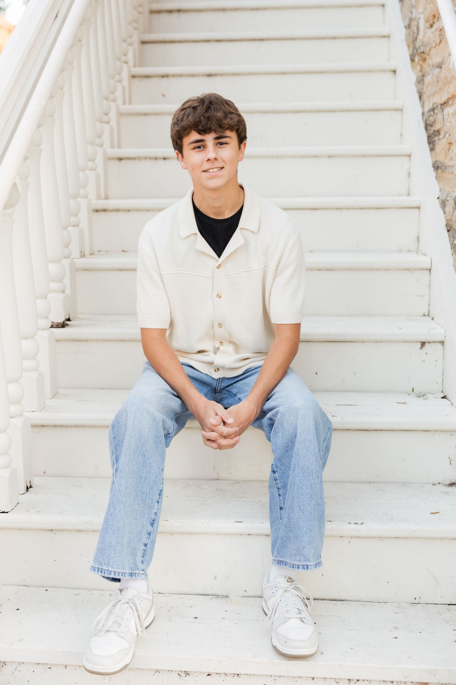 Teen boy in cream shirt and jeans sitting on white outdoor stairs, smiling at camera.