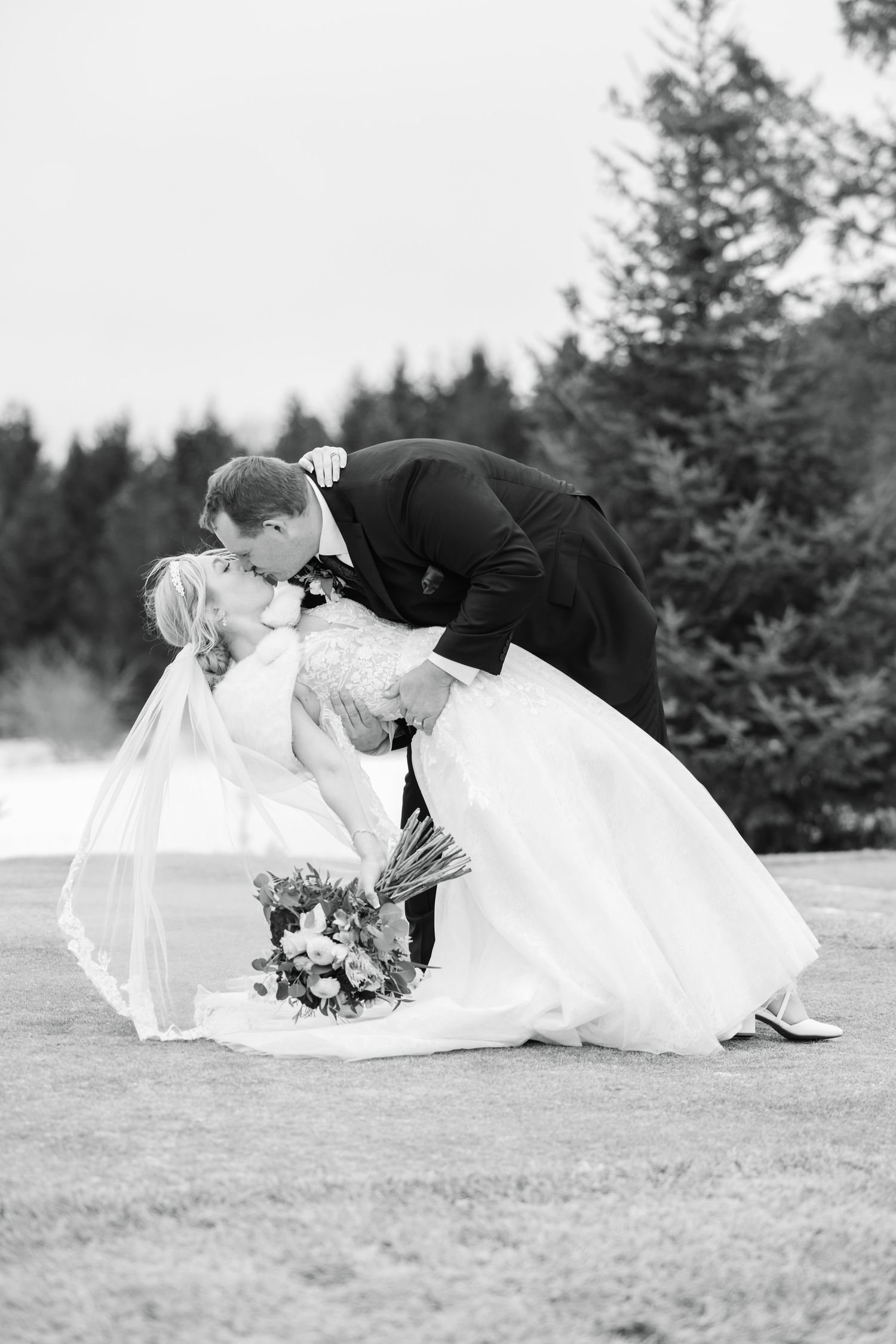 Romantic wedding moment captured in black and white on a grassy field with evergreen trees.