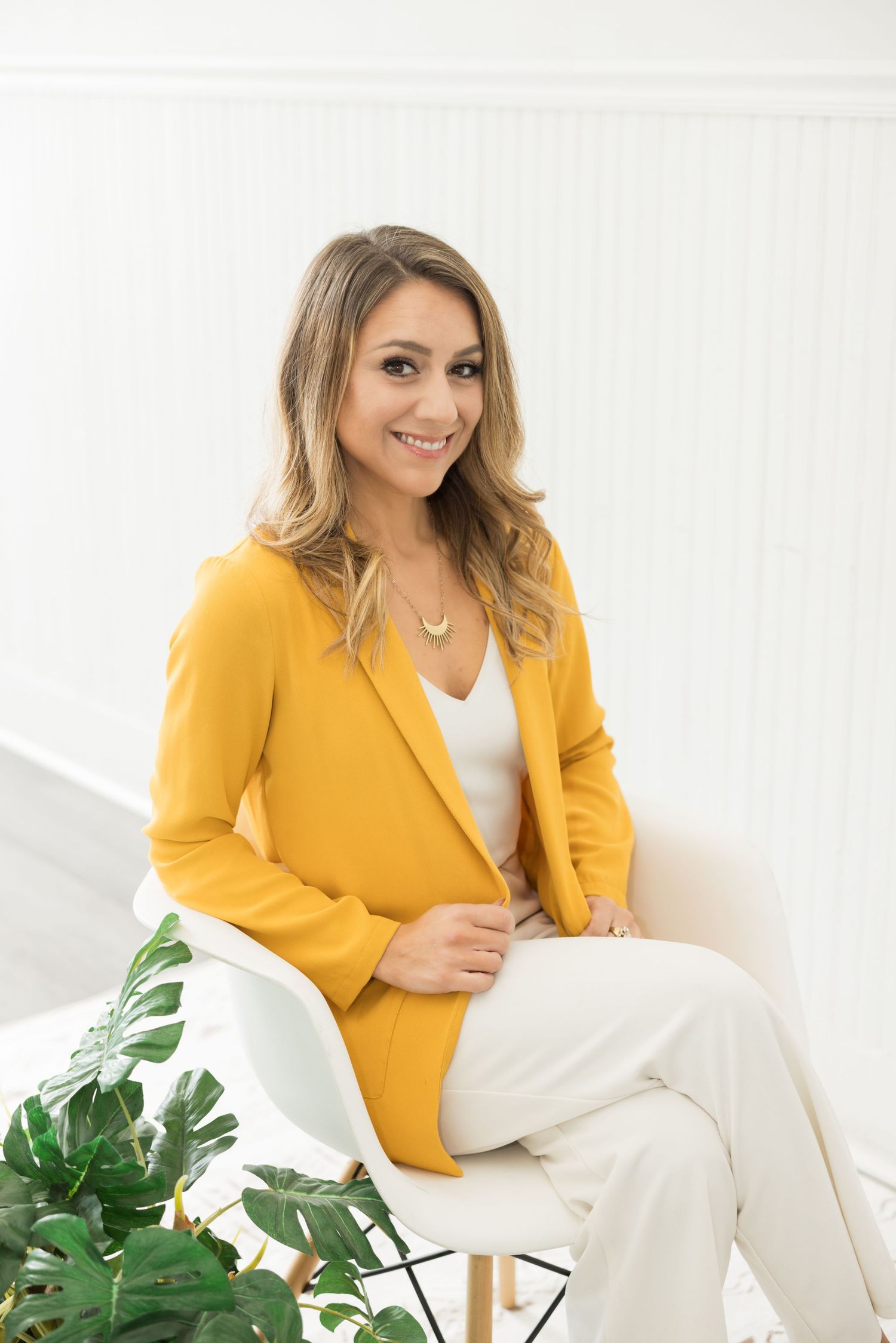 Professional wearing a yellow blazer and white pants sits elegantly in a modern office setting with green plants.