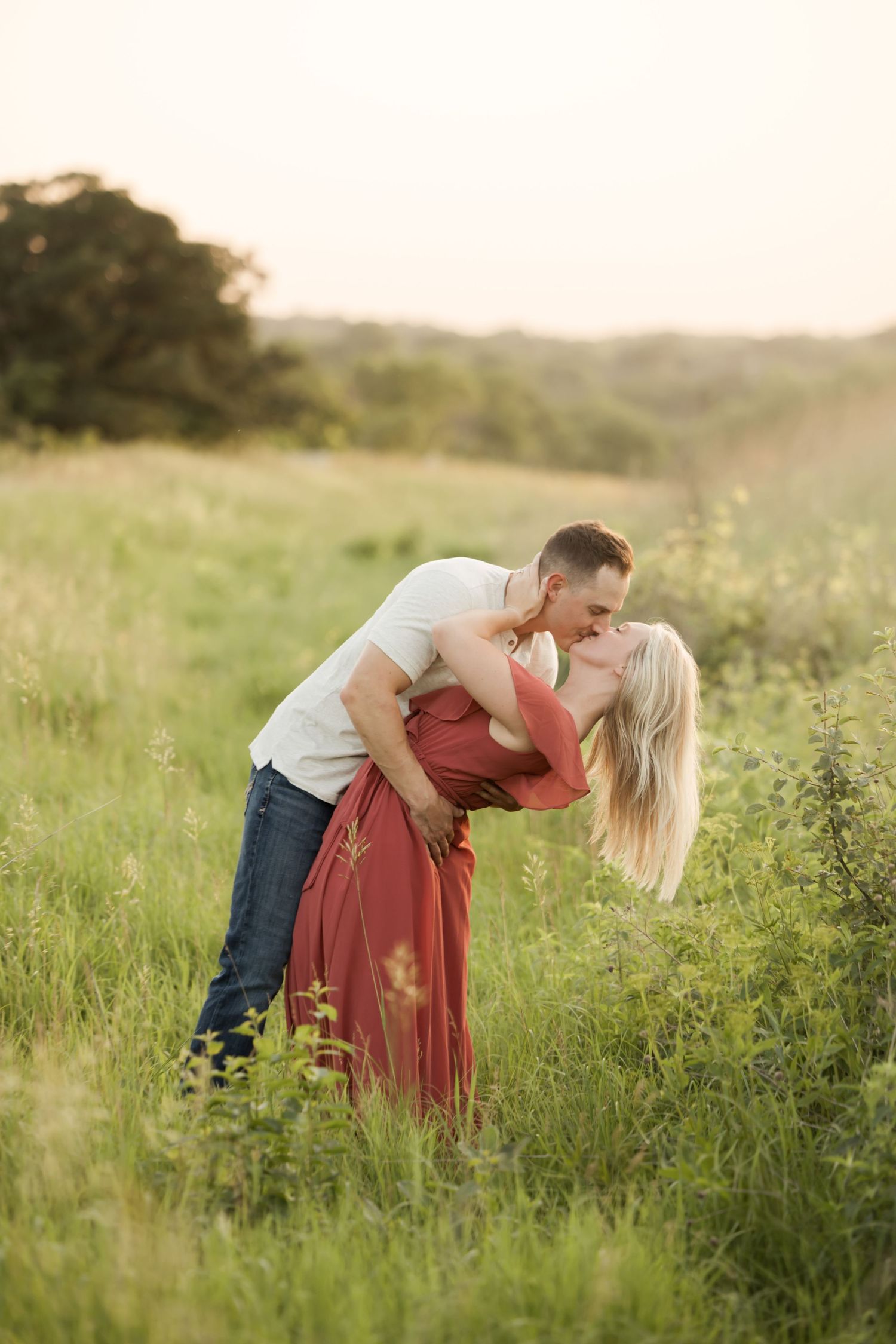 A romantic couple embraces in a sunlit meadow with tall grass and wildflowers during golden hour.