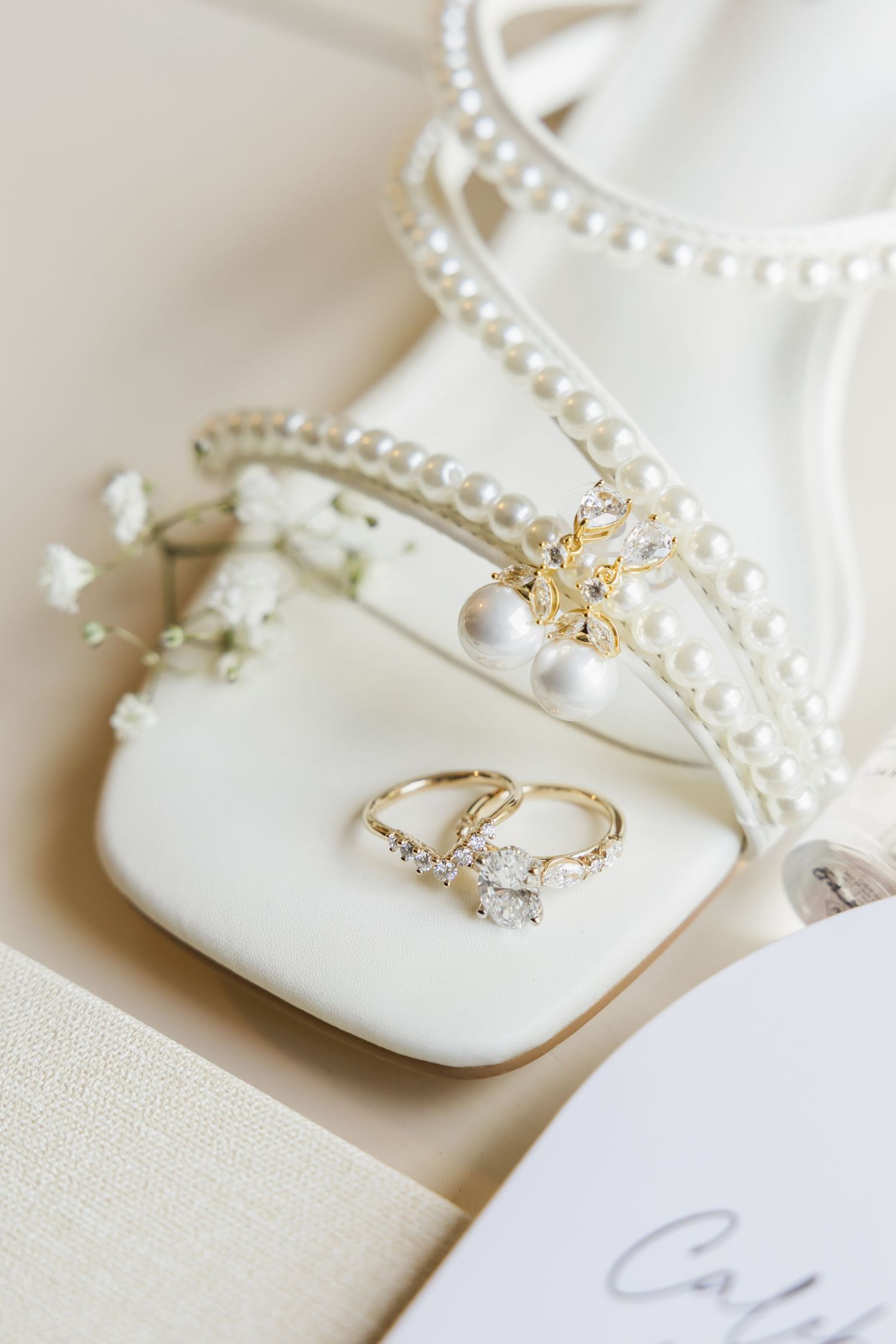 Wedding details featuring pearl-adorned shoes and diamond engagement rings on white fabric.