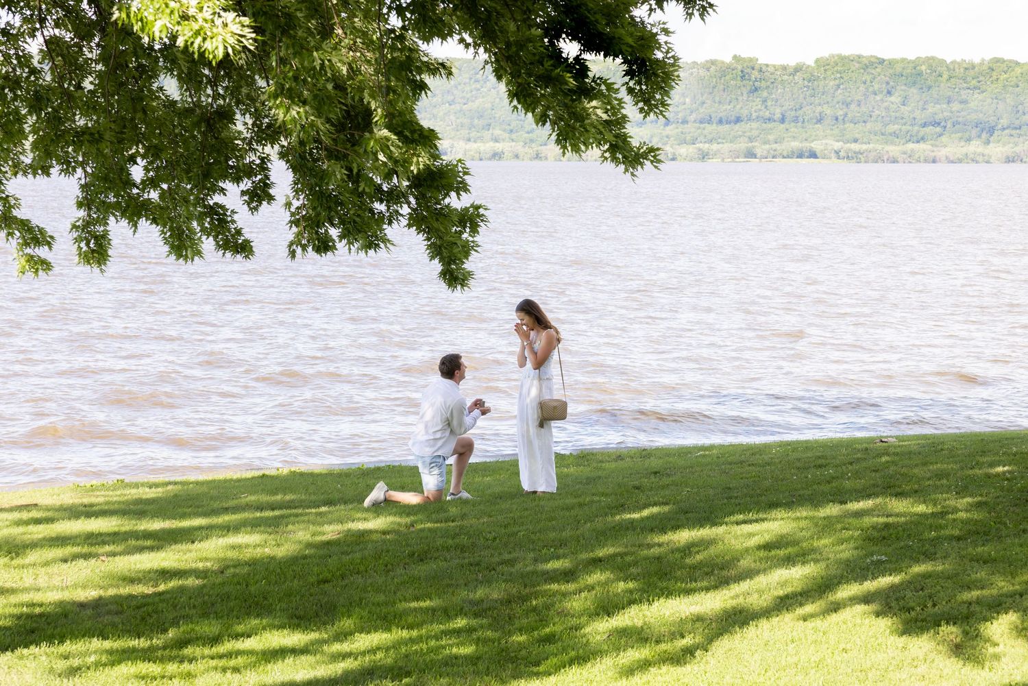 Two people in white outfits sit on a grassy slope by a lake enjoying a peaceful summer afternoon.