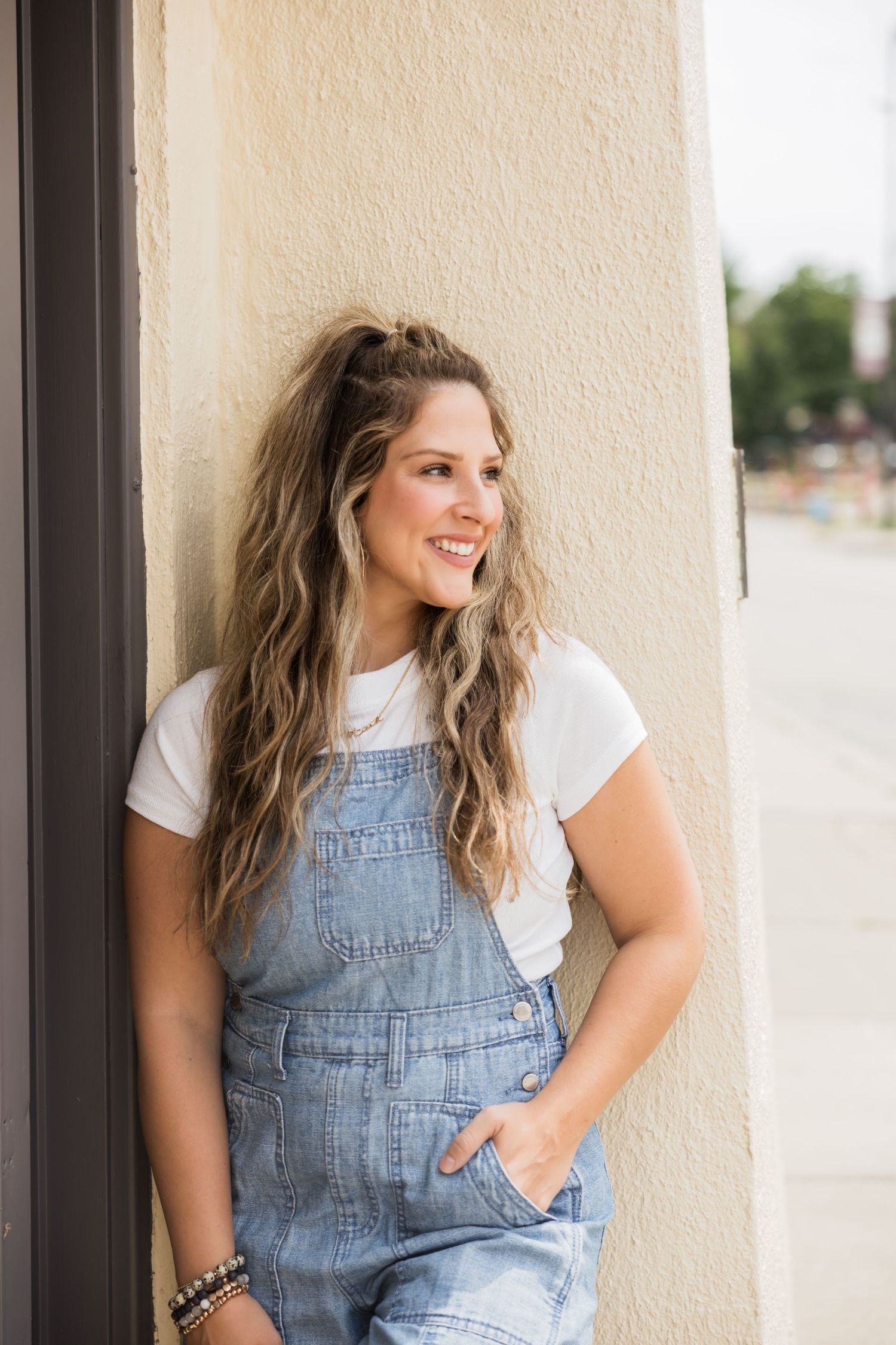 Woman in denim overalls and white t-shirt smiles while leaning against a white wall outdoors.