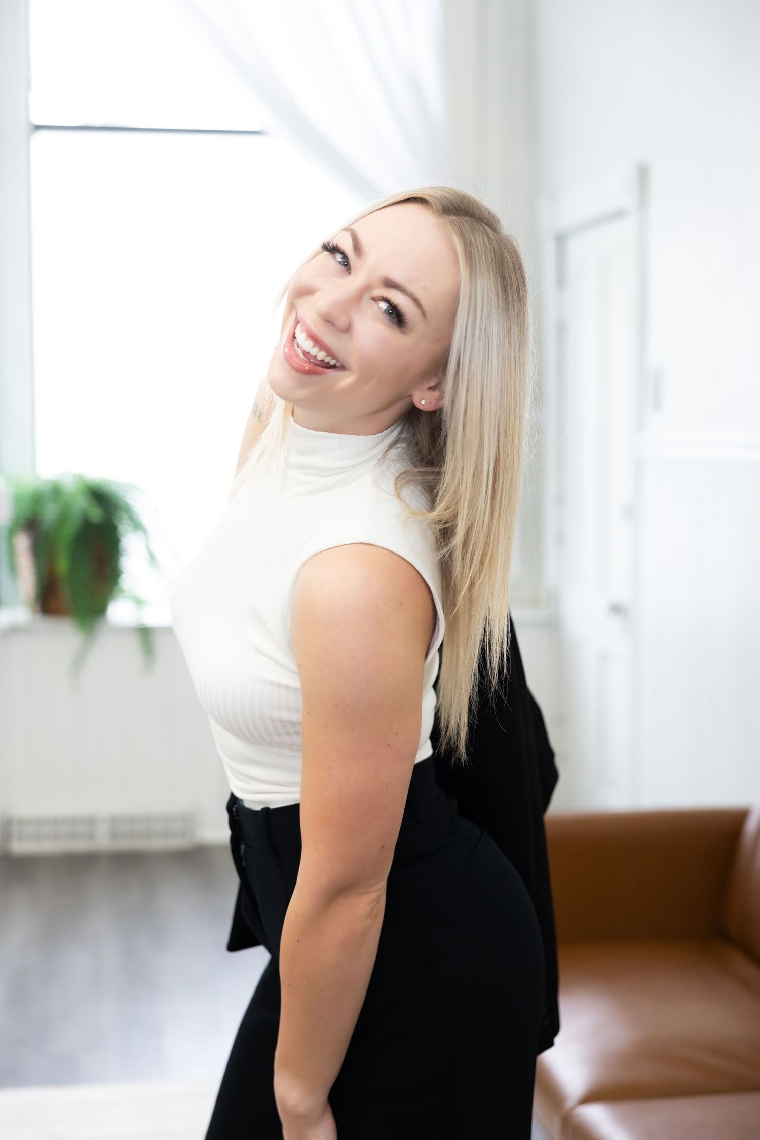 A blonde woman in a white sleeveless top laughs joyfully while looking up in a bright interior setting.