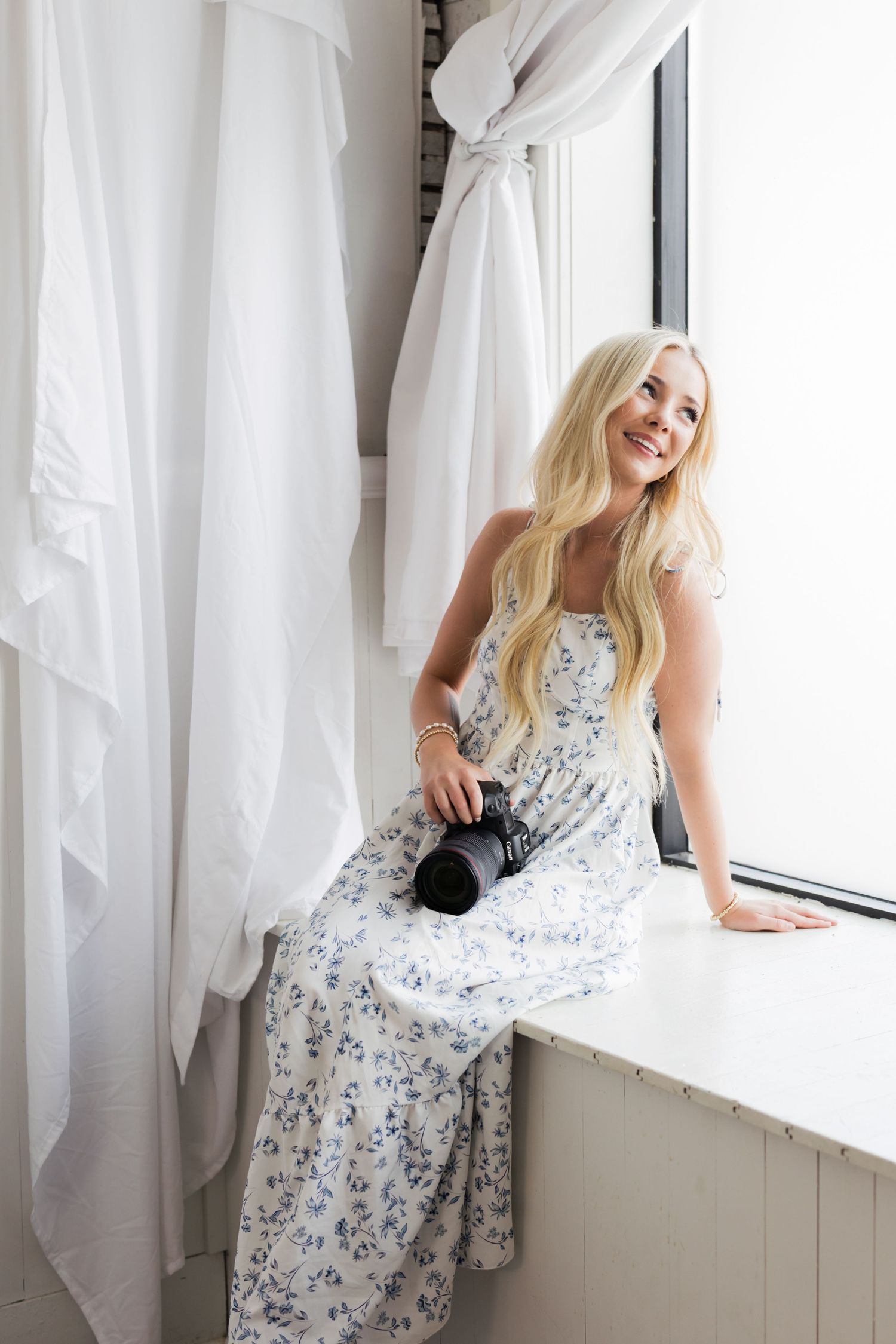Smiling blonde woman in floral maxi dress holding a camera while sitting on a white windowsill with white curtains.