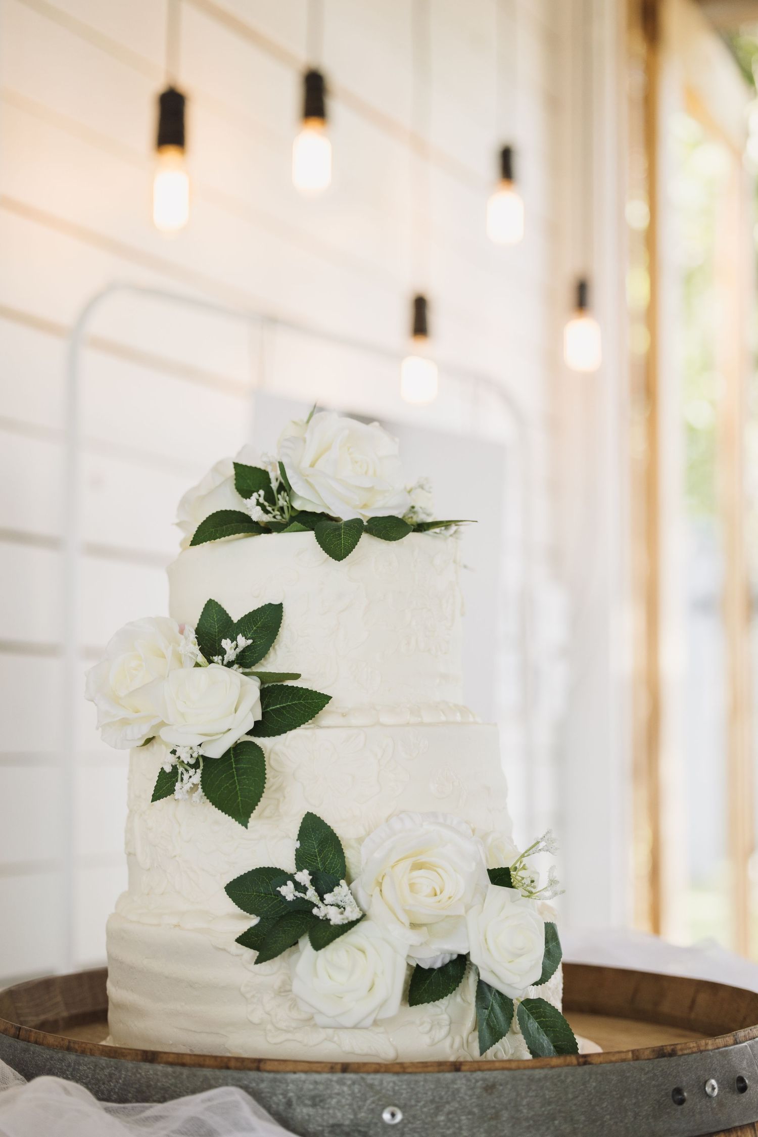 Simple white wedding cake adorned with fresh roses and dark green leaves against a light background.