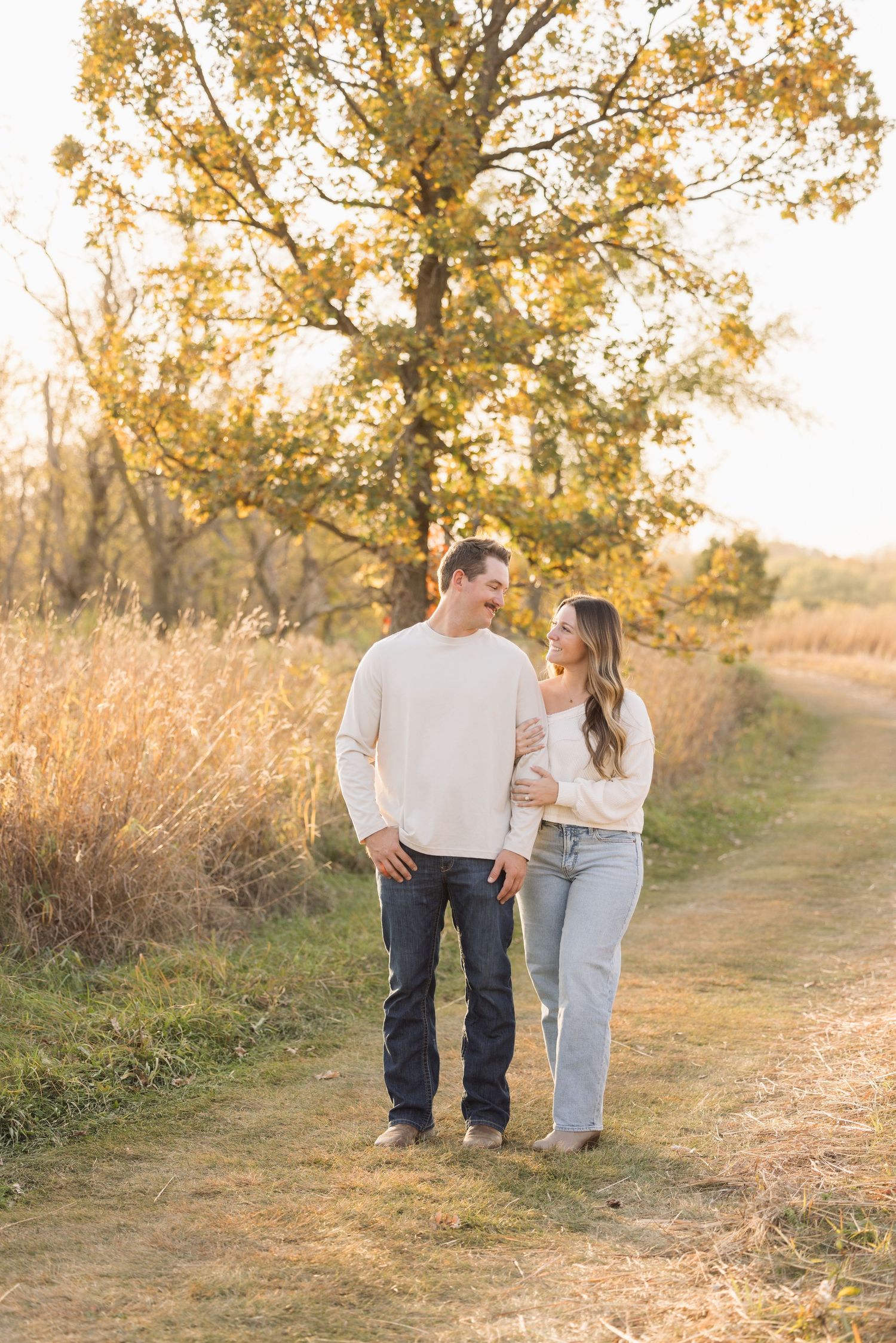 A couple walks together on a dirt path surrounded by golden autumn trees and tall grass at sunset.