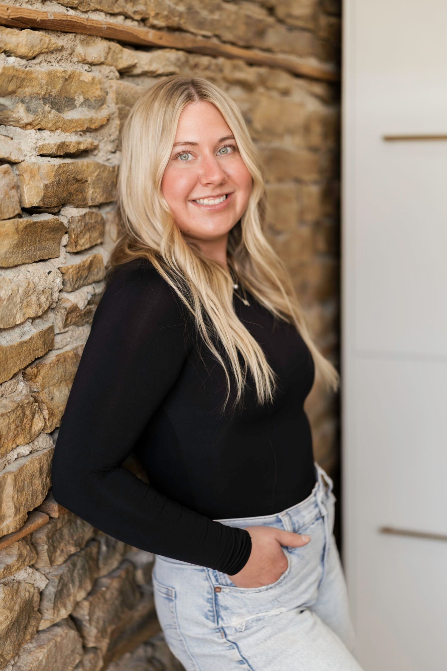 A person in a black sweater and light wash jeans poses against a rustic stone wall with a warm smile.