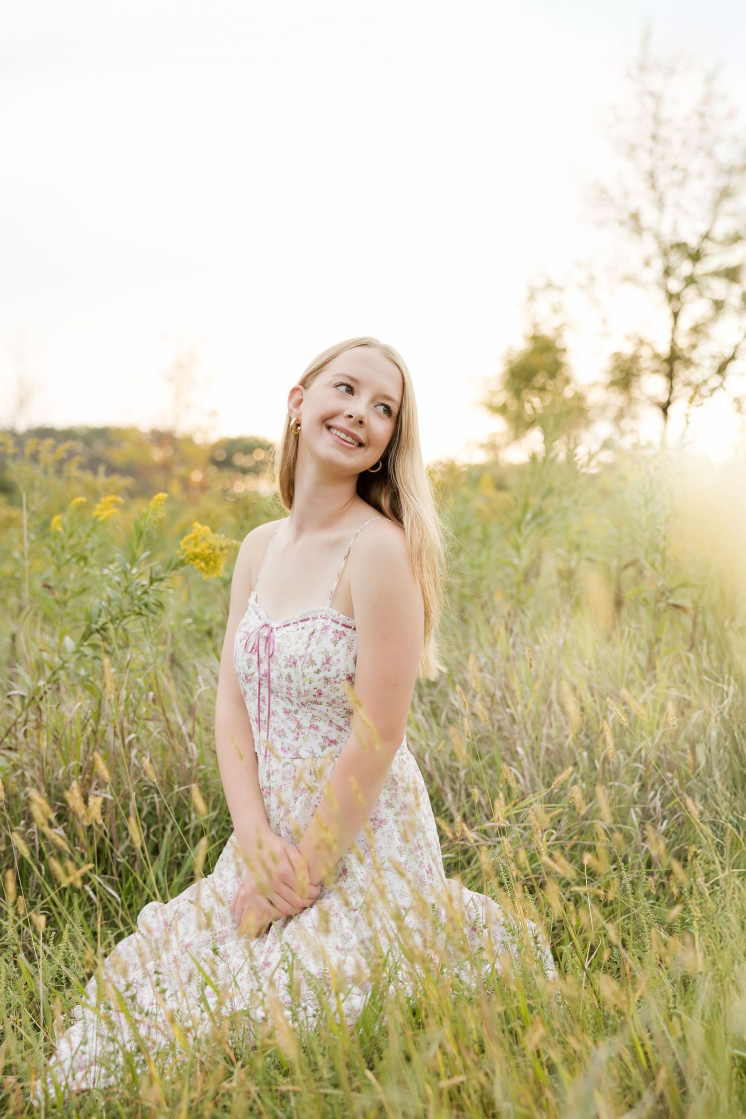 Young blonde woman in floral sundress sitting in golden meadow, smiling upward in warm sunset light.