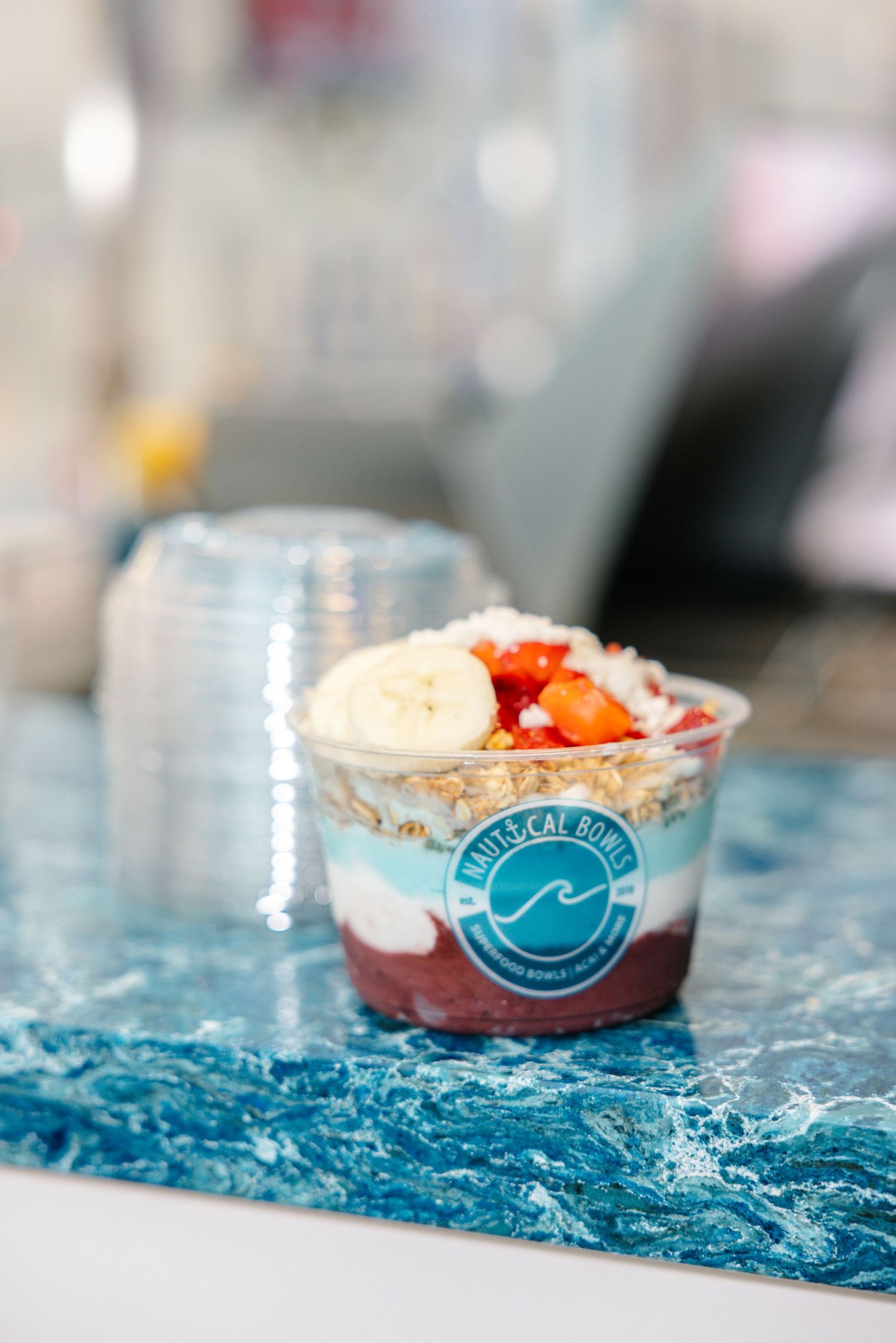A clear plastic cup filled with acai bowl topped with fresh fruit and granola sits on a blue marble surface.