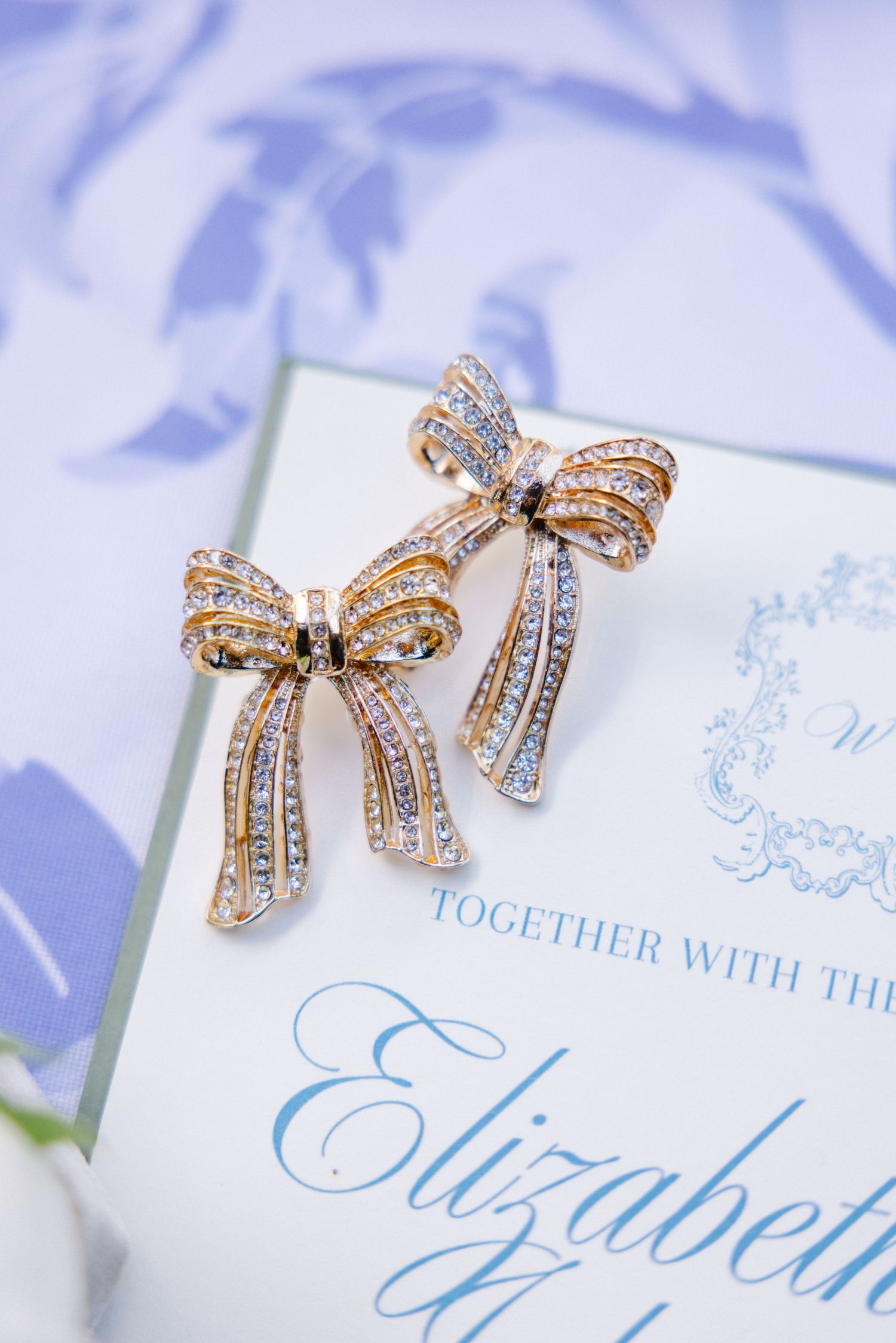 Gold bow-shaped crystal earrings on elegant wedding invitation with blue calligraphy.