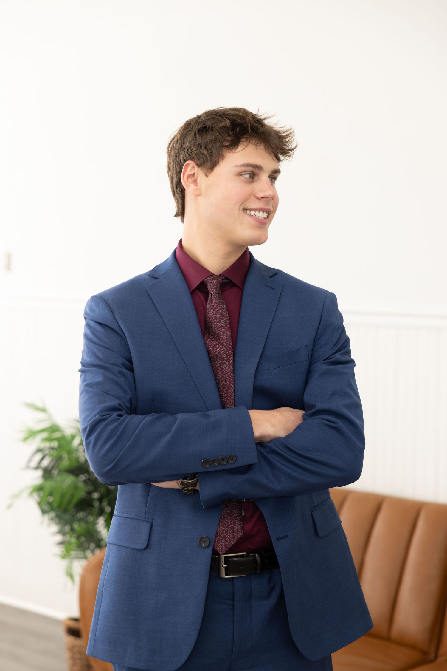 Professional in navy suit and burgundy shirt poses against white wall with brown couch and plant in background.