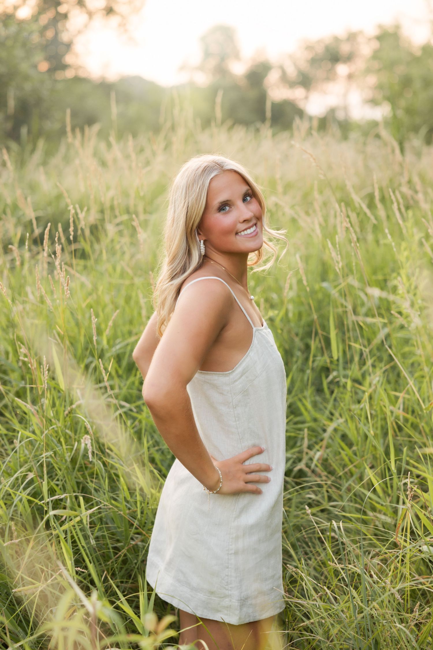 Smiling blonde woman in white sundress standing in tall green grass field during golden hour sunset.