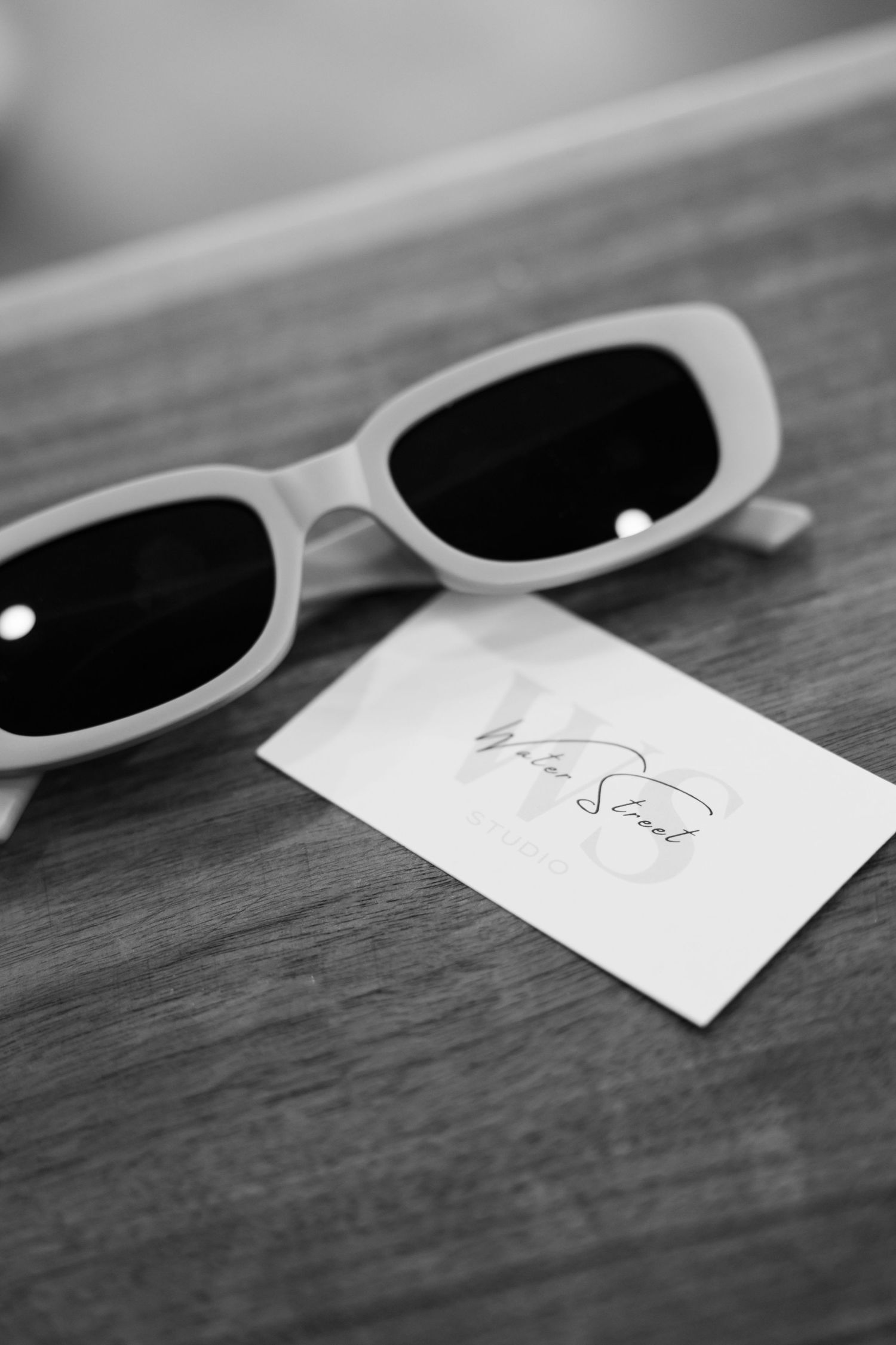 A pair of white sunglasses with dark lenses sits next to a small piece of paper with a signature on a wooden surface.