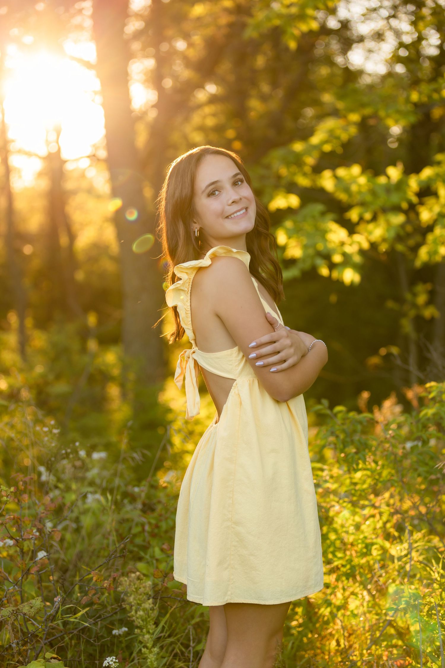 Young woman in yellow dress smiling outdoors during golden sunset in lush green forest.