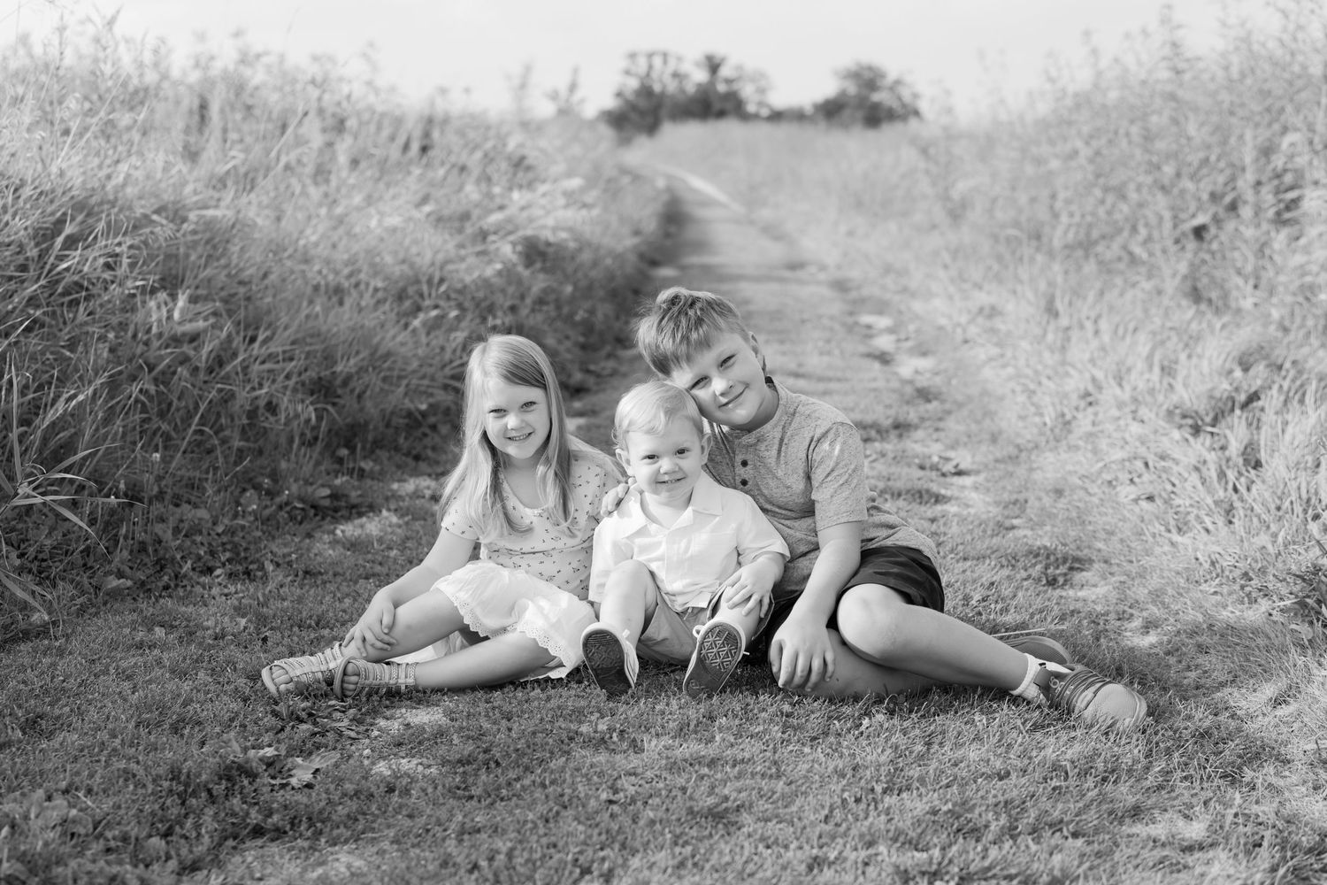 Black and white photo of three children sitting together on a rural dirt path surrounded by tall grass.