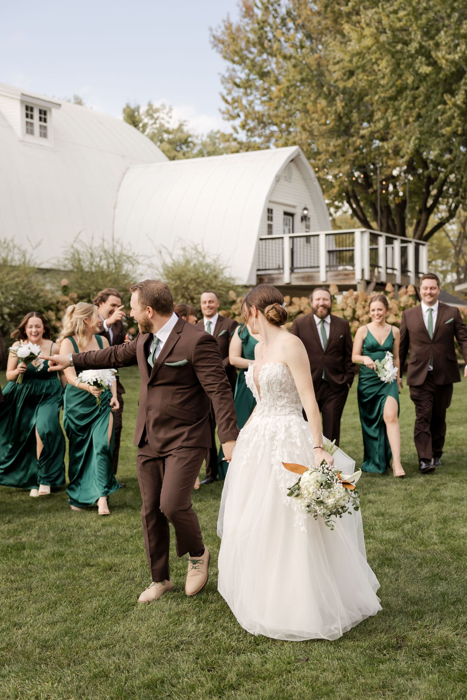 Wedding party in brown suits and emerald green dresses poses playfully on a grassy lawn.