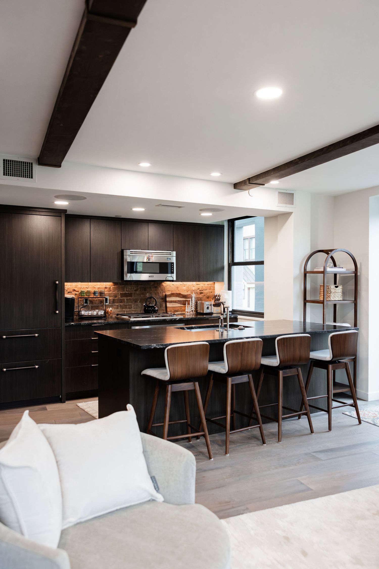 Modern open-concept kitchen with dark cabinetry, brick backsplash, black island with bar stools, and exposed ceiling beams.